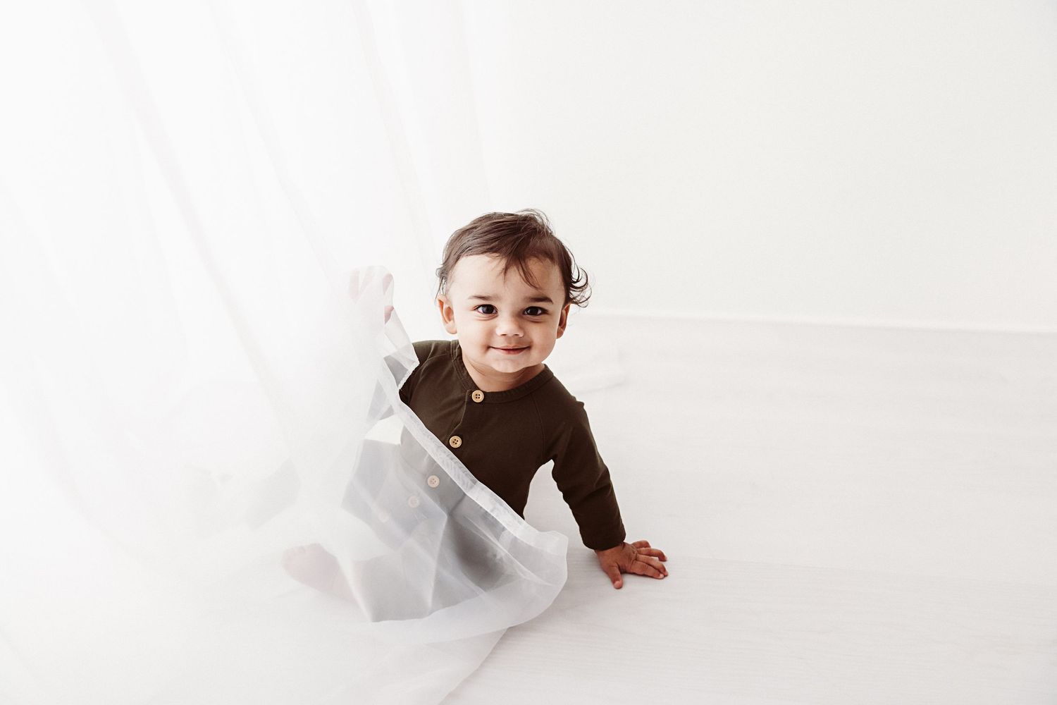 Happy baby crawling on a white surface with sheer curtains in a bright studio.