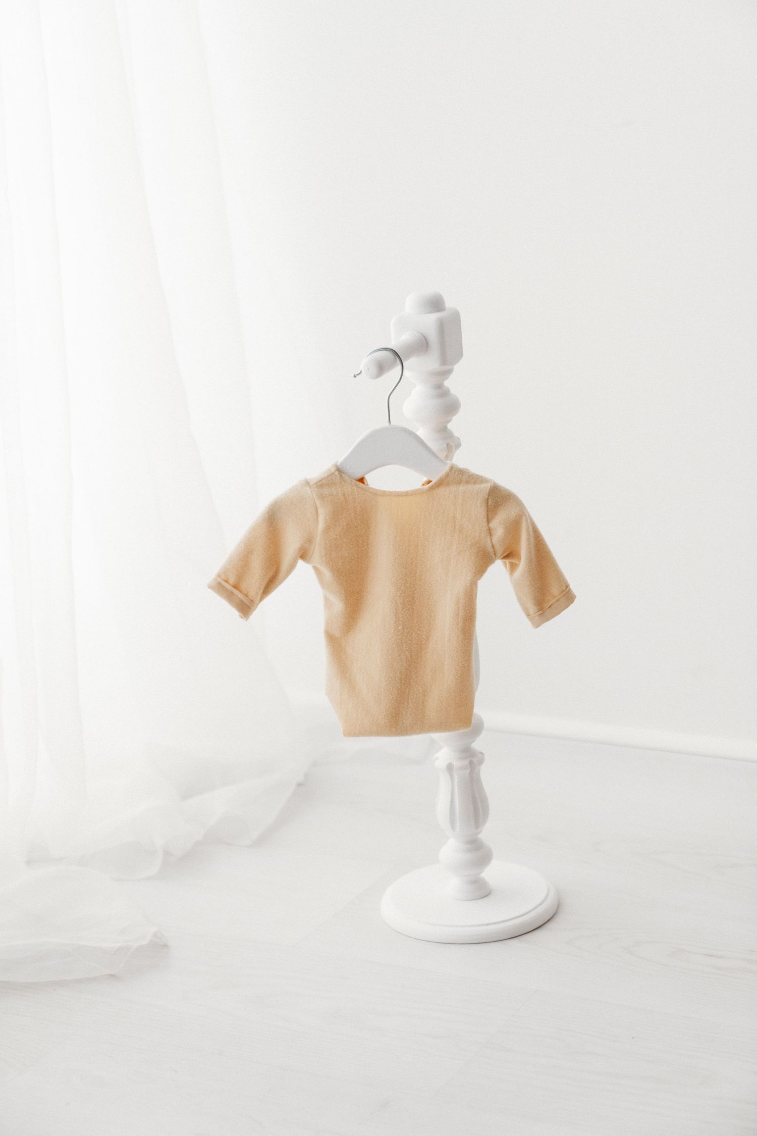 Tan ribbed baby bodysuit with short sleeves hanging on a white decorative stand in a bright white room.