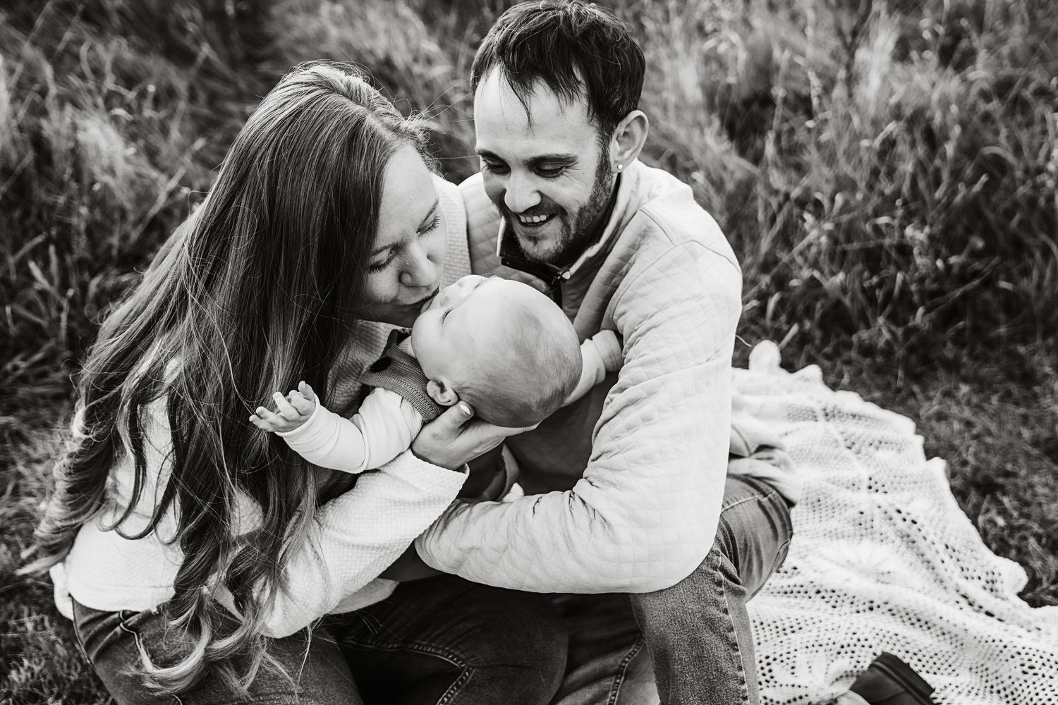 Black and white photo of happy parents laughing with their baby on a blanket.