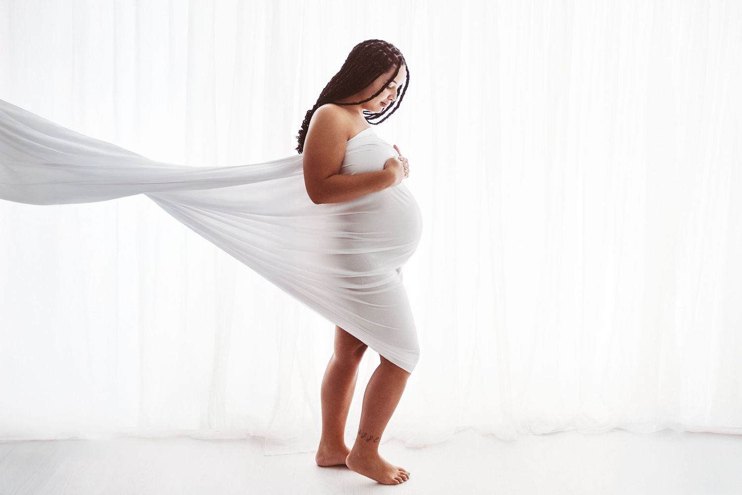 Pregnant woman wrapped in flowing white fabric, cradling her baby bump in a bright white studio setting.