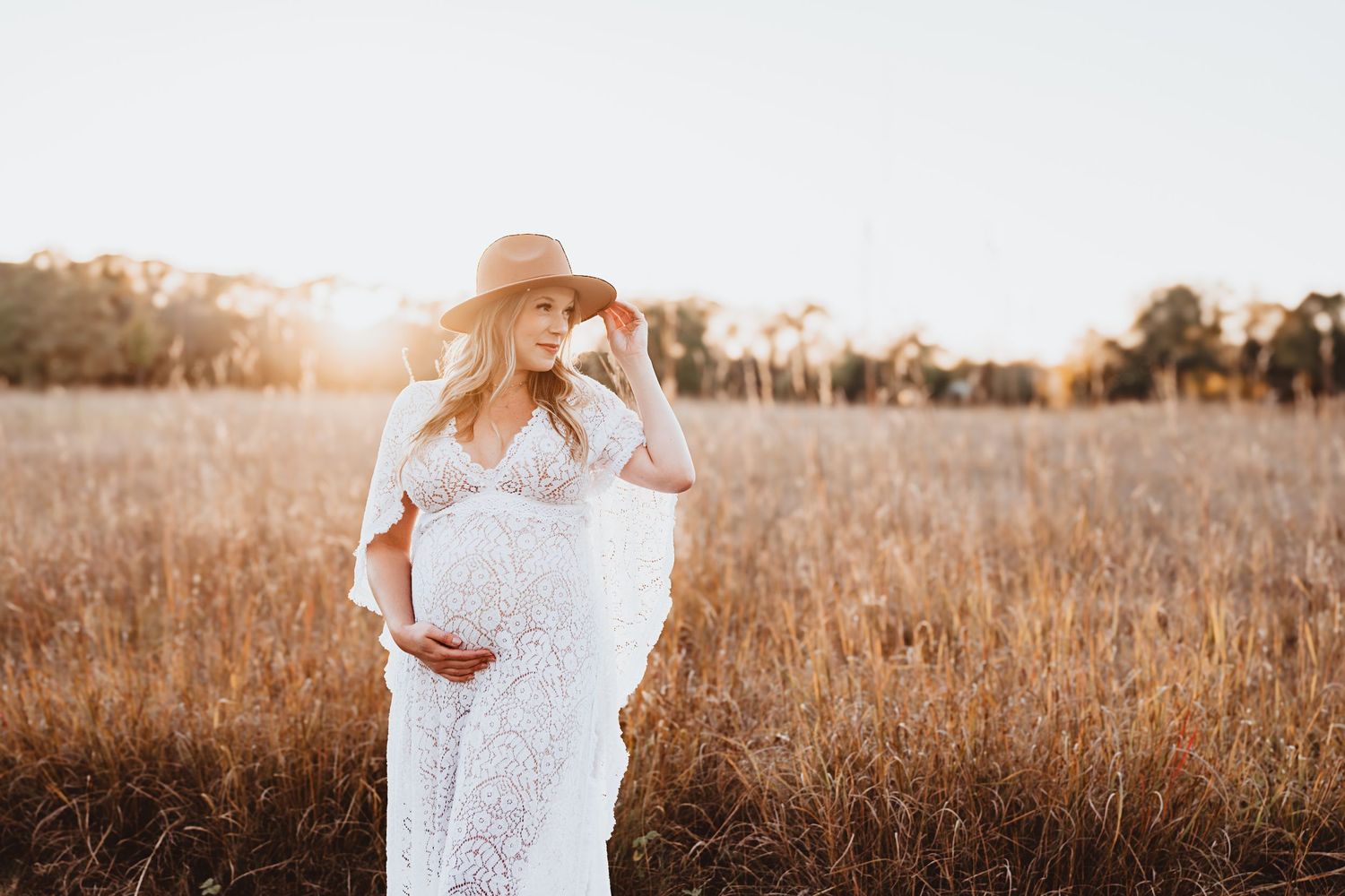 Pregnant woman in white lace dress and sun hat standing in a golden wheat field at sunset.