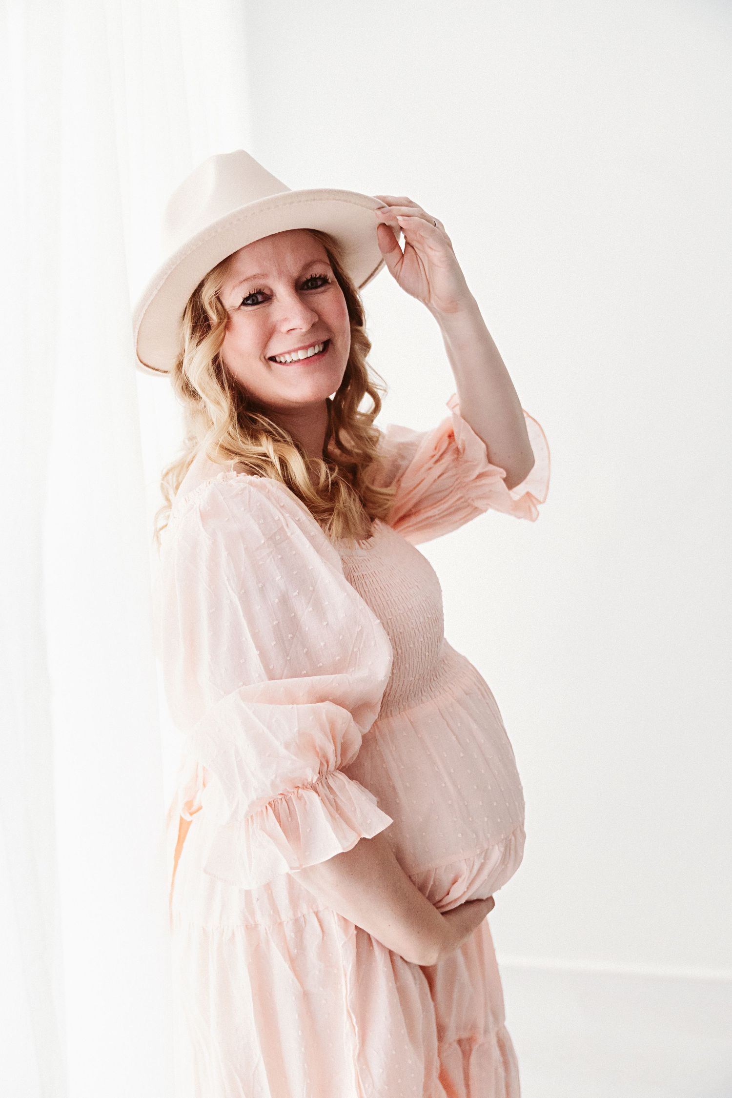 Smiling pregnant woman in pink dress and white hat holding baby bump in bright white studio.