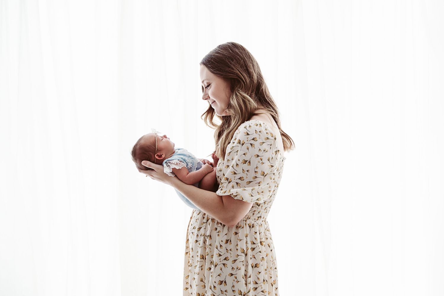 Young mother in floral dress holding her newborn baby against a bright white curtain backdrop.