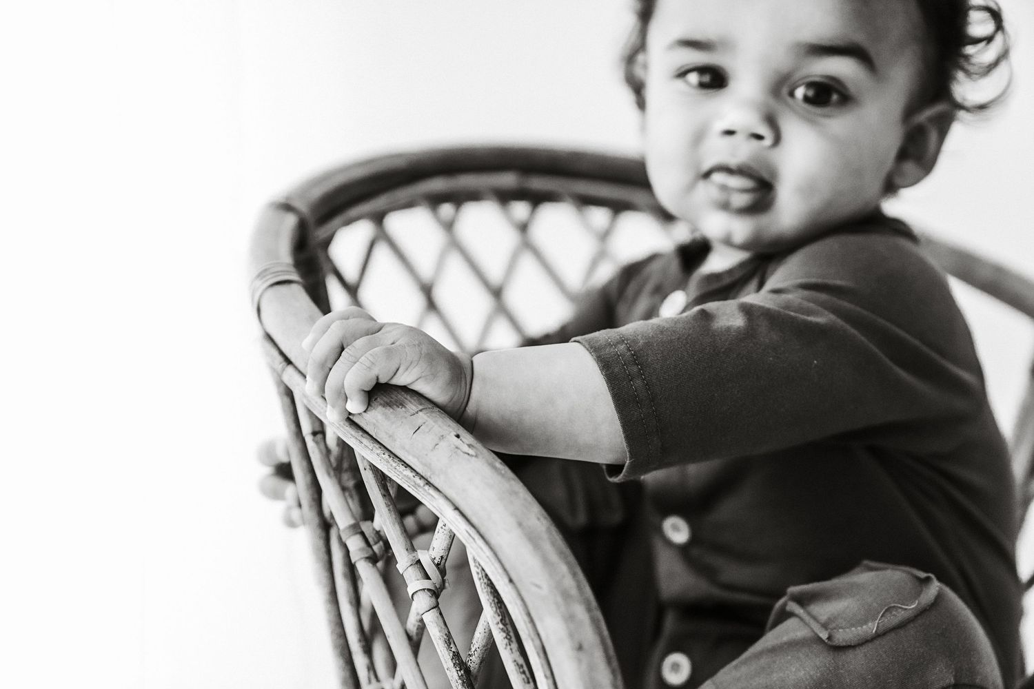 Black and white photo of a toddler leaning on a wicker chair, looking at the camera.