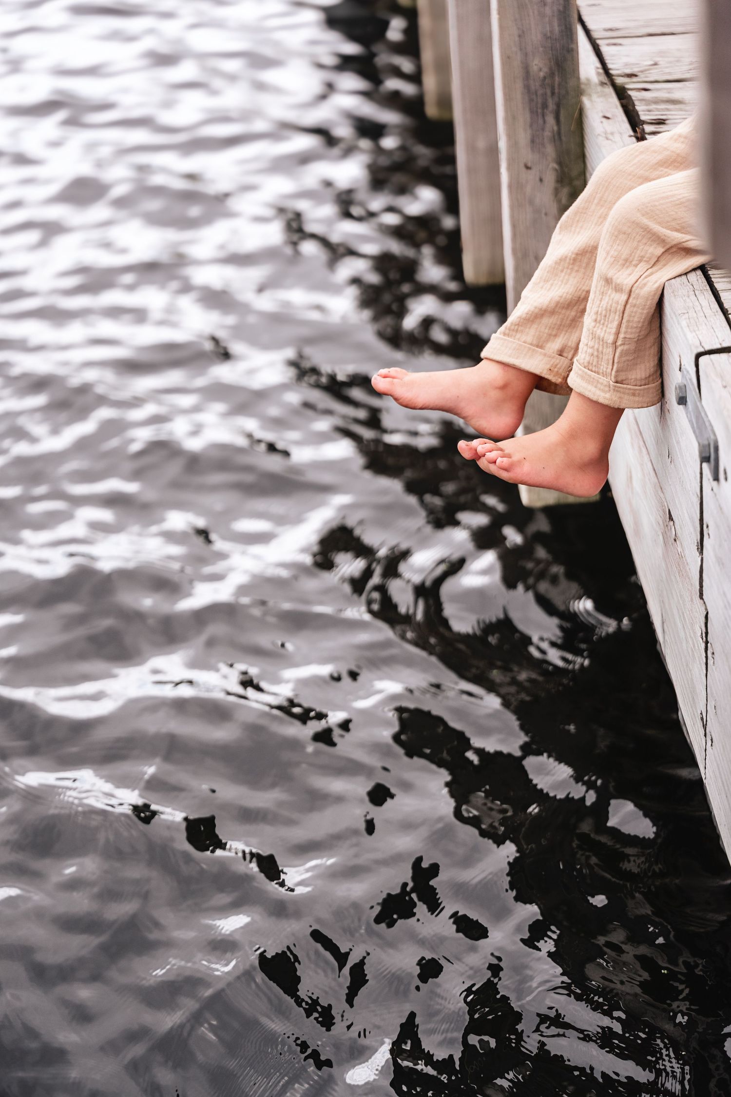 Barefoot child dangling feet over wooden pier above rippling water.