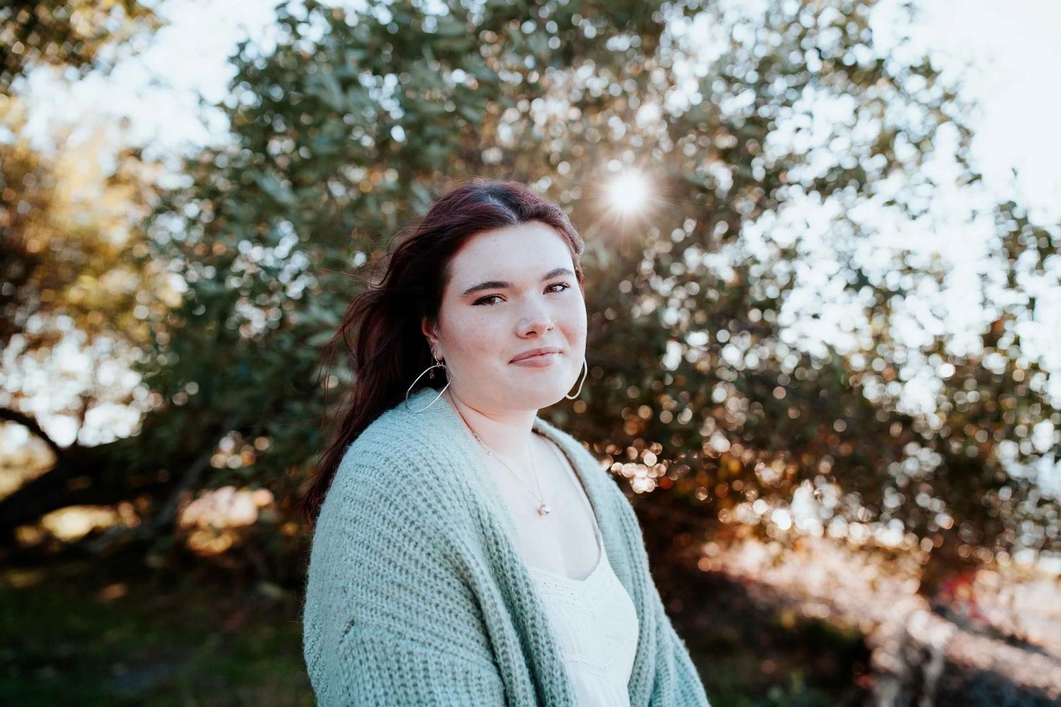 Young woman in mint green cardigan smiling outdoors with sunlight filtering through trees behind her.