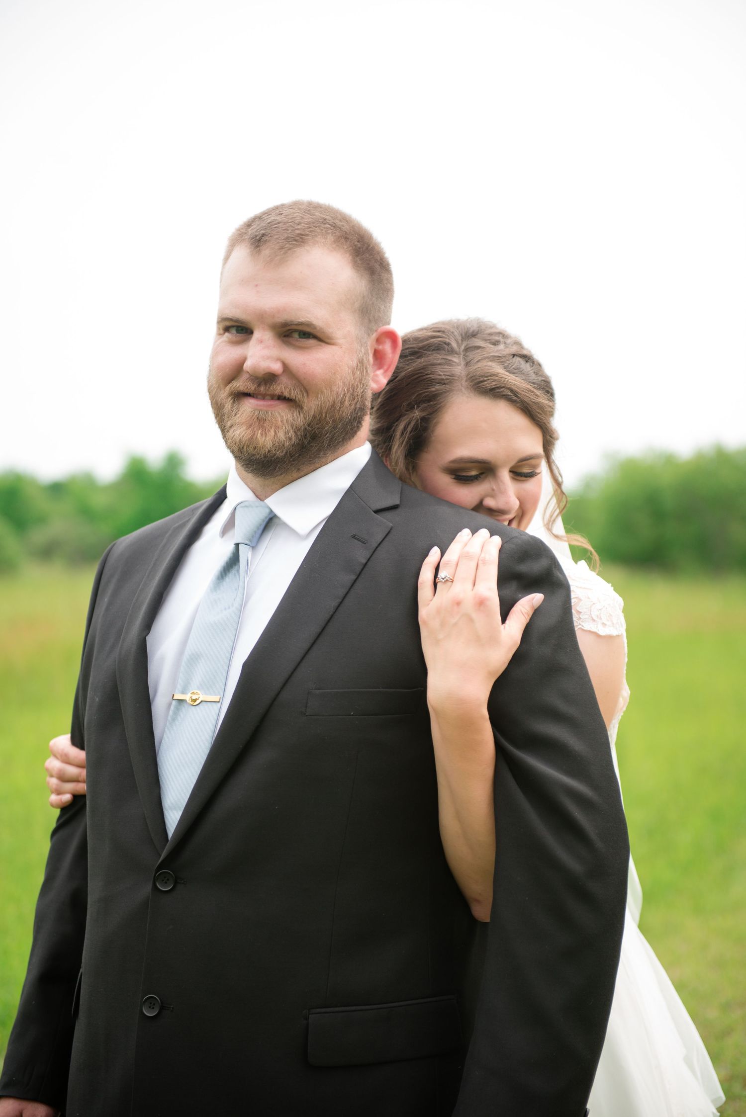 Bride resting her head on groom's shoulder in a green field, showing off her diamond wedding ring.