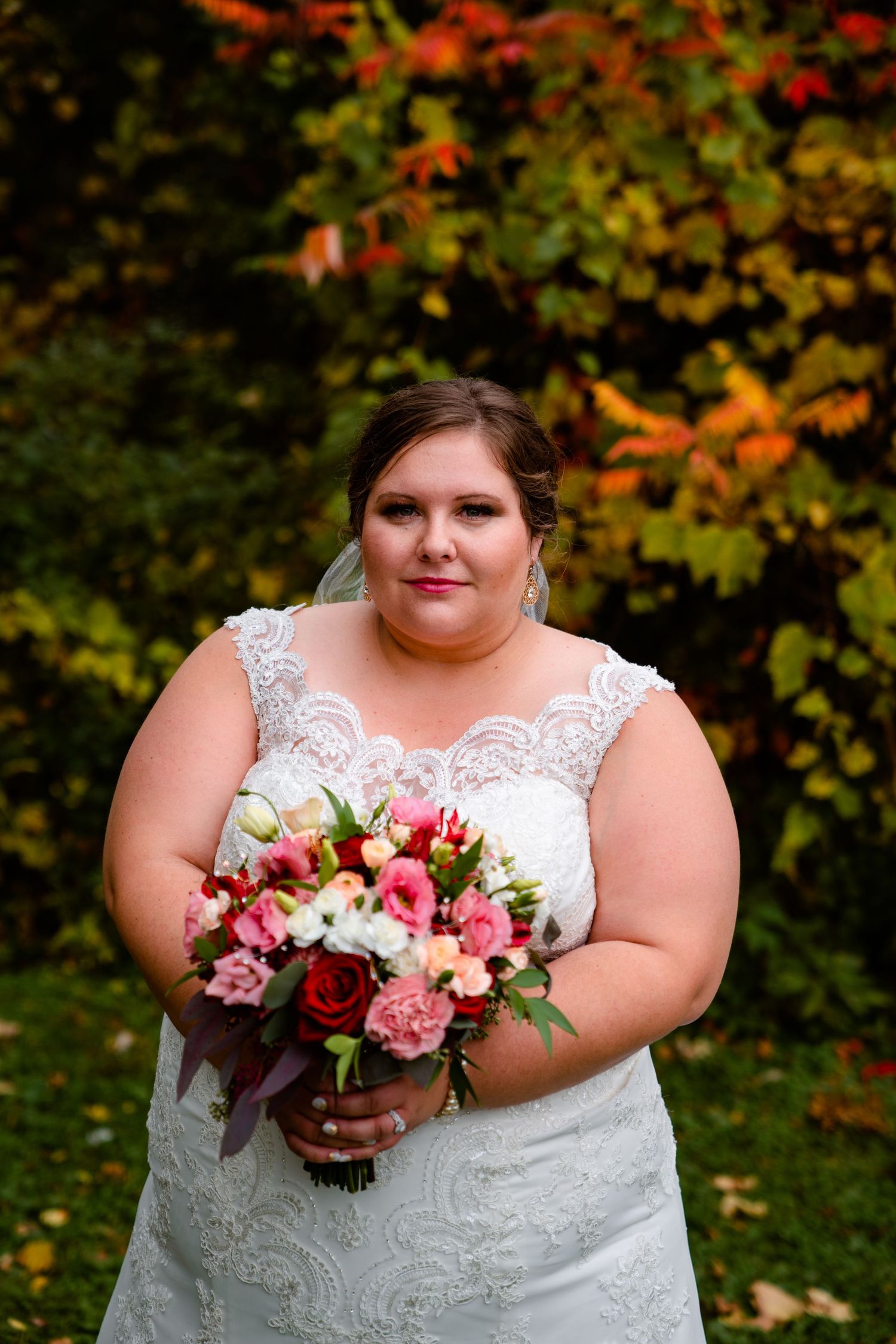 Plus-size bride in lace gown holds pink and red bouquet before colorful autumn foliage backdrop.