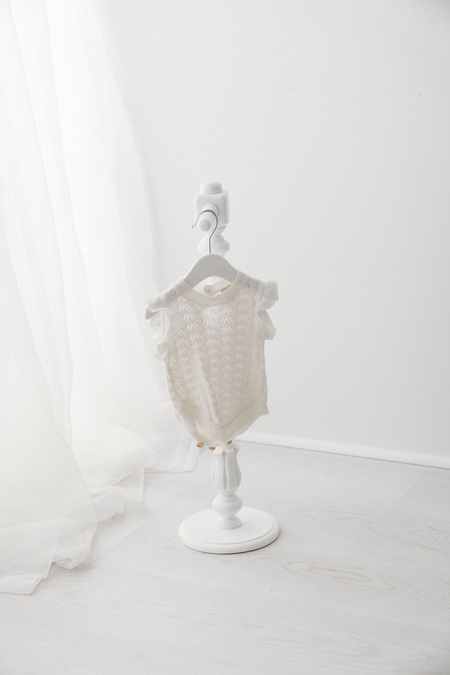 Delicate cream knit baby onesie with ruffled trim displayed on a white decorative mini stand.