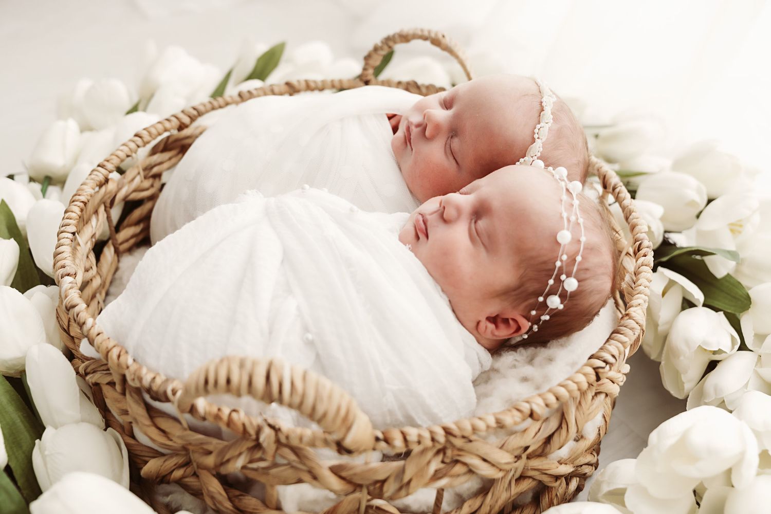 Two sleeping newborn twins wrapped in white swaddles resting in a basket with white tulips.