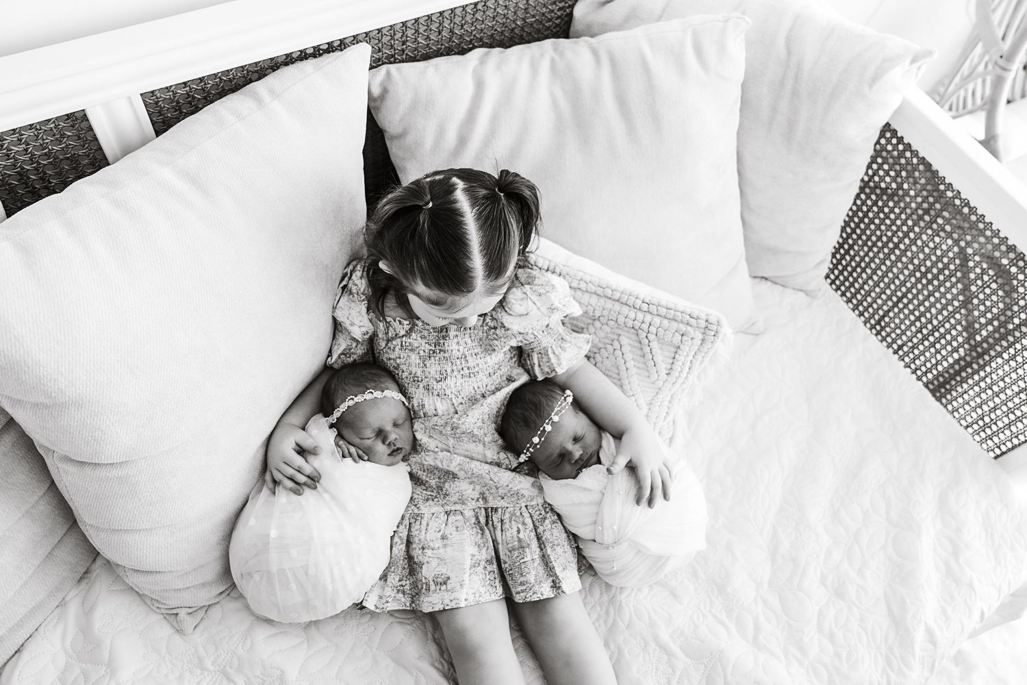 Black and white photo of young girl cuddling twin newborn babies on a white bed.