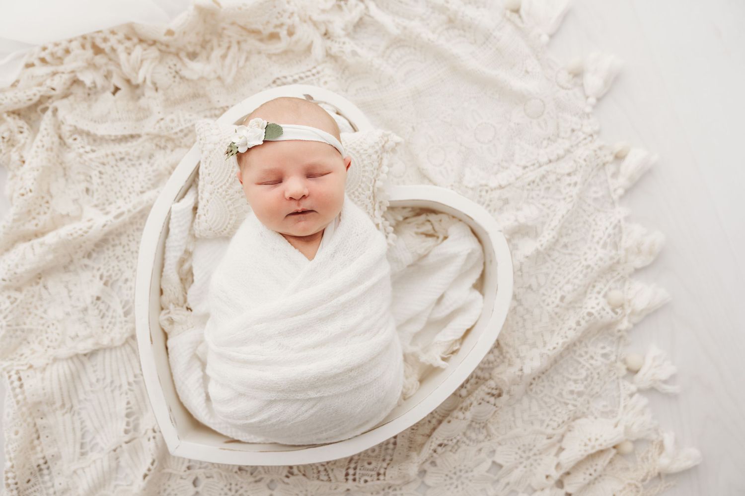 Swaddled newborn baby girl with floral headband sleeping in a heart-shaped white bowl prop.