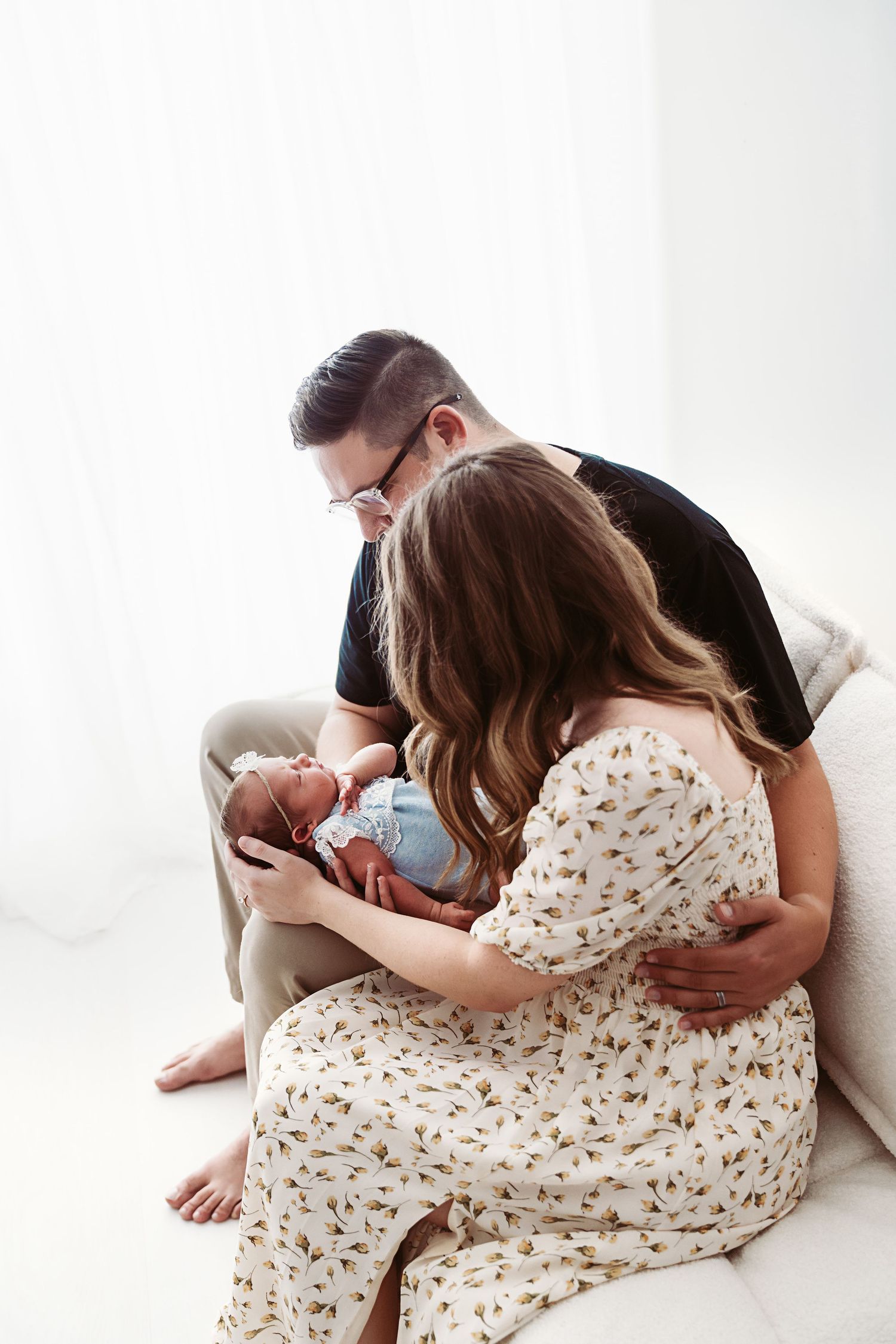 New parents sitting on a couch lovingly gazing at their newborn baby in a bright studio.