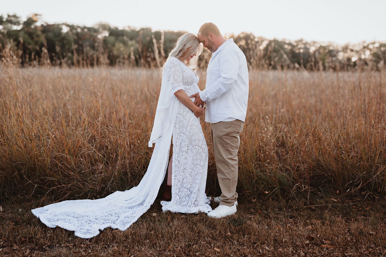 Couple sharing tender moment in field at sunset, woman in white lace maternity gown.