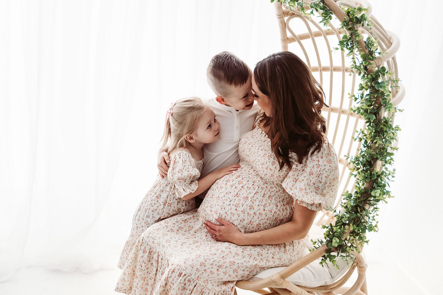 Pregnant mother sitting in rattan swing chair sharing tender moment with two young children.