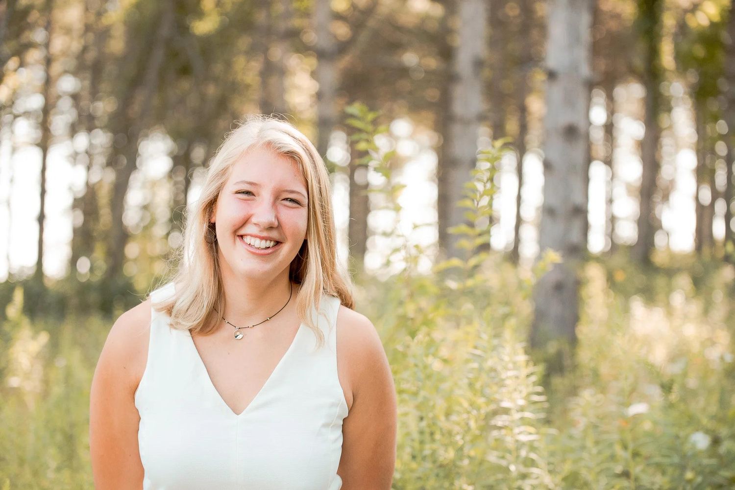 Smiling blonde woman in white top standing outdoors among tall trees and wildflowers in warm golden light.