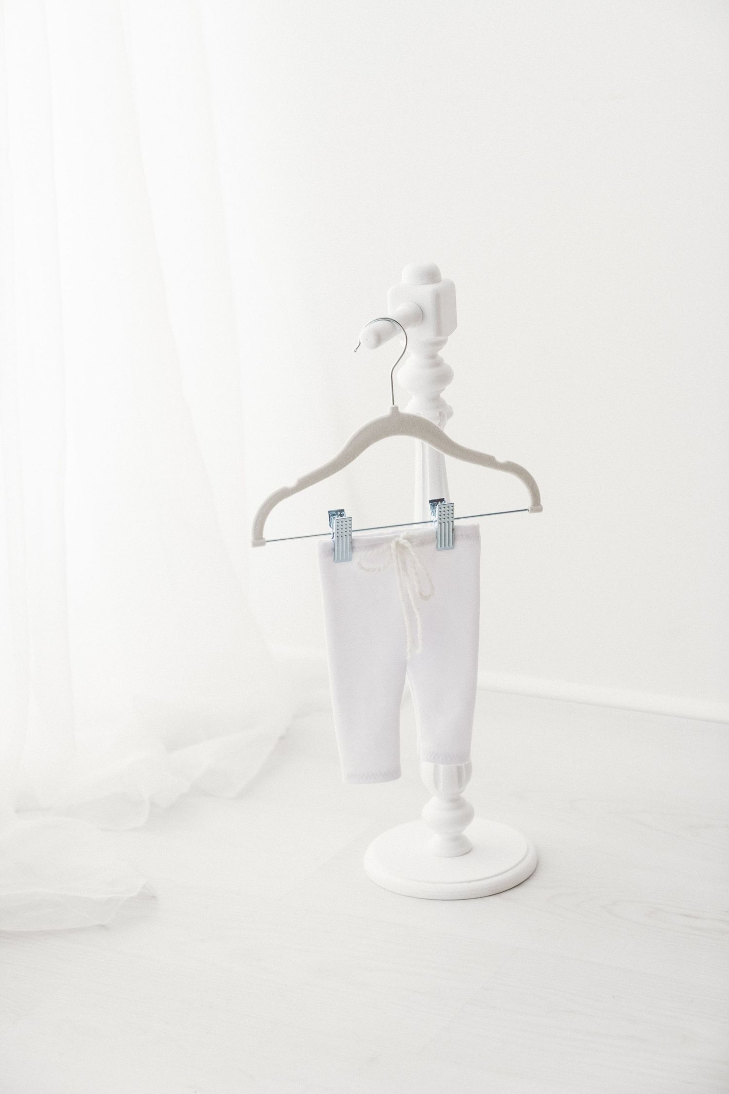 White baby pants with drawstring waist clipped to a velvet hanger on a white decorative stand.