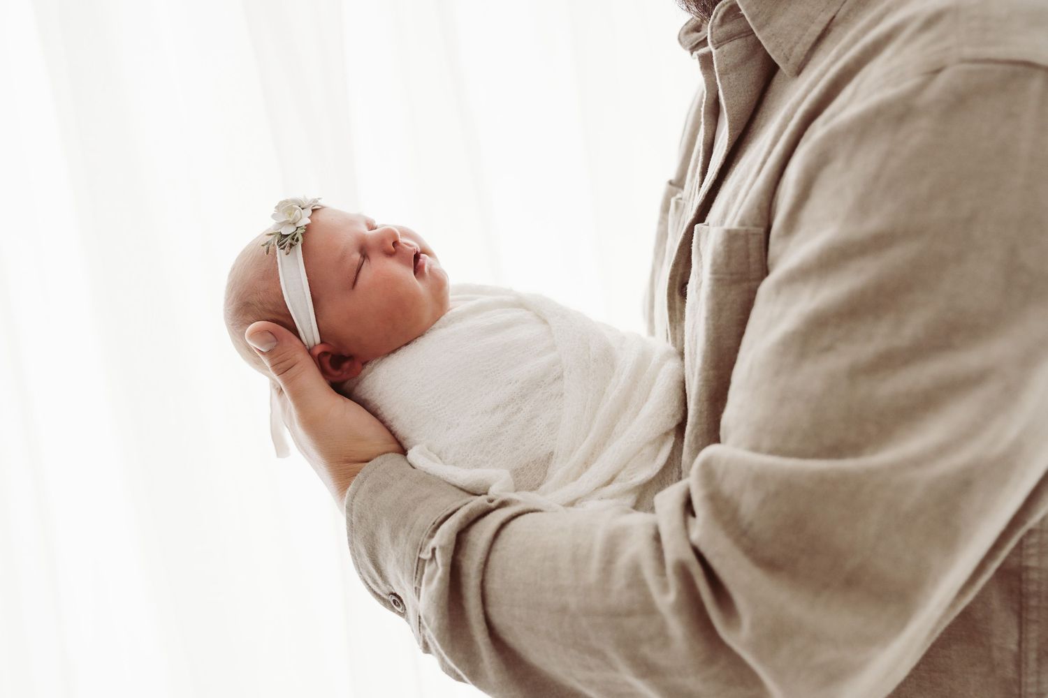 Close-up of a newborn baby girl swaddled in white with a floral headband, cradled in fathers arms.