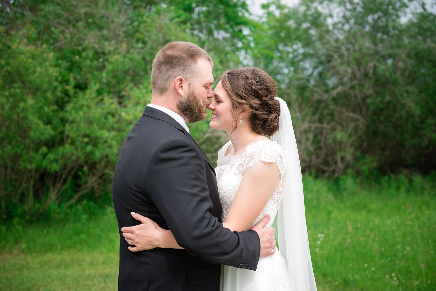 Bride and groom sharing an intimate almost-kiss moment surrounded by lush green trees outdoors.