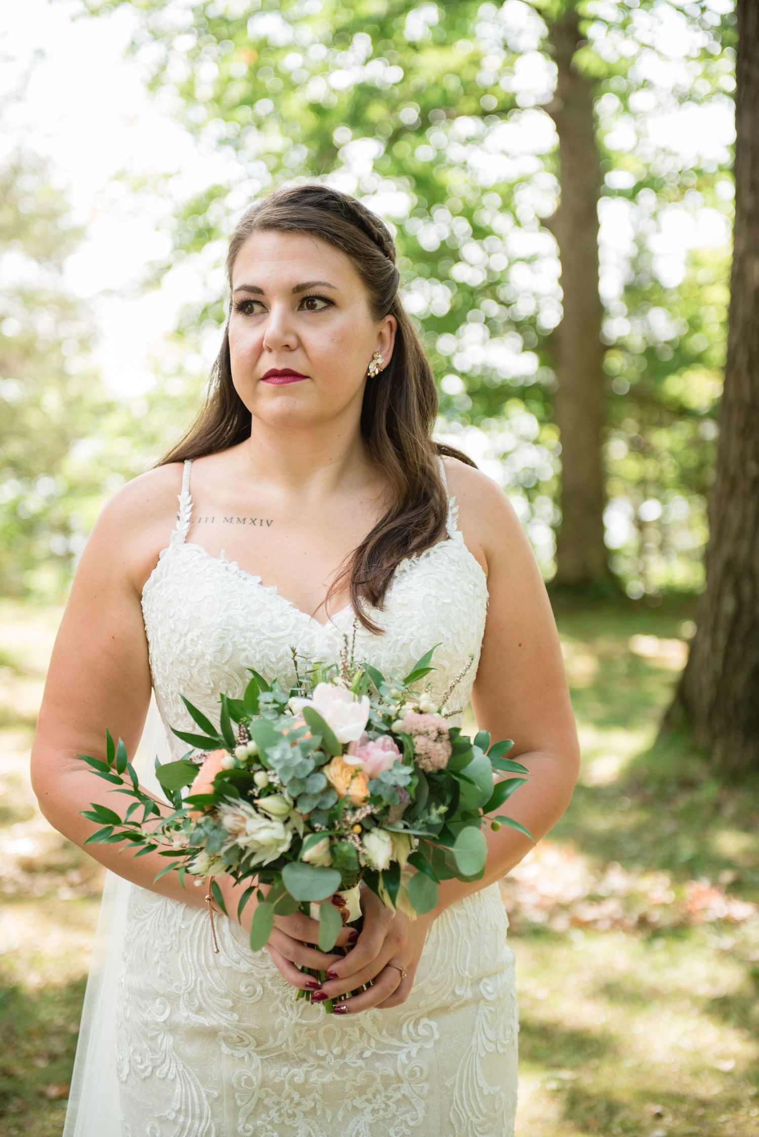 Bride in lace spaghetti strap gown holding bouquet of roses and eucalyptus in wooded setting.