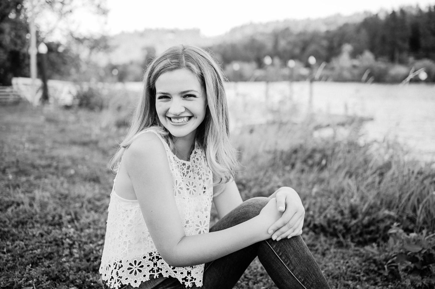Smiling young woman with long blonde hair sitting in grass outdoors in a black and white portrait photo.