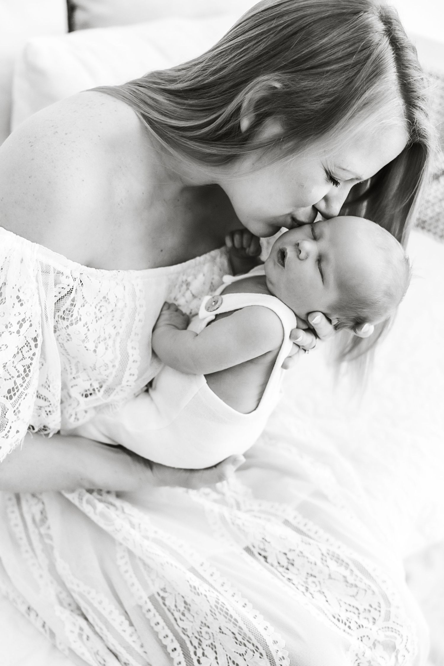 Black and white portrait of a mother in lace dress tenderly kissing her newborn baby.