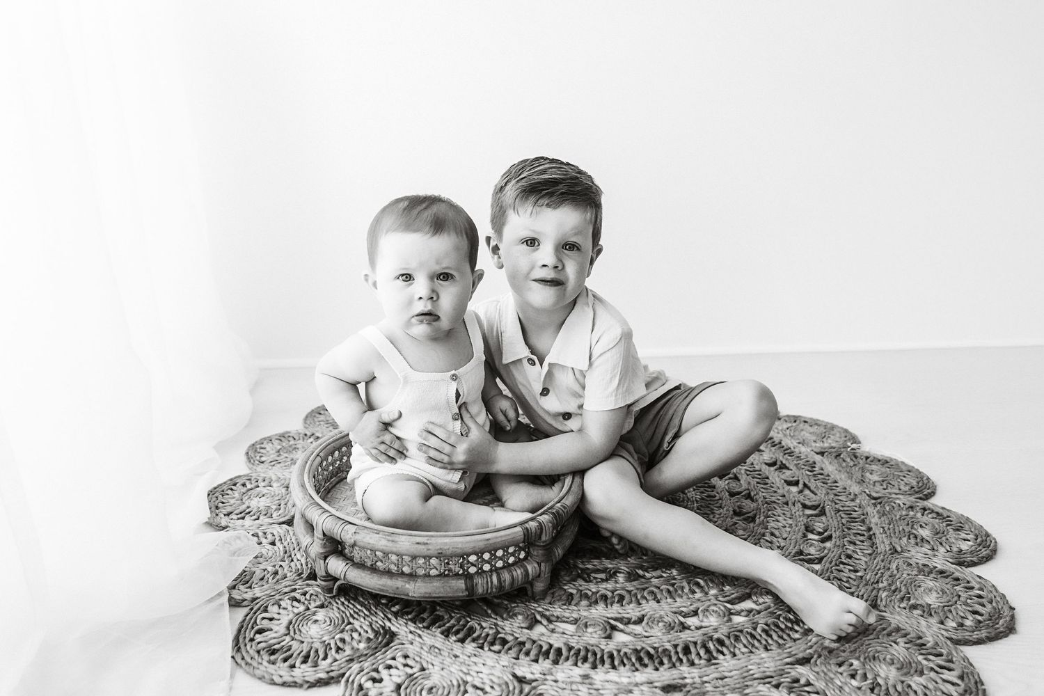 Two young brothers pose together on a woven rug in a black and white studio portrait.