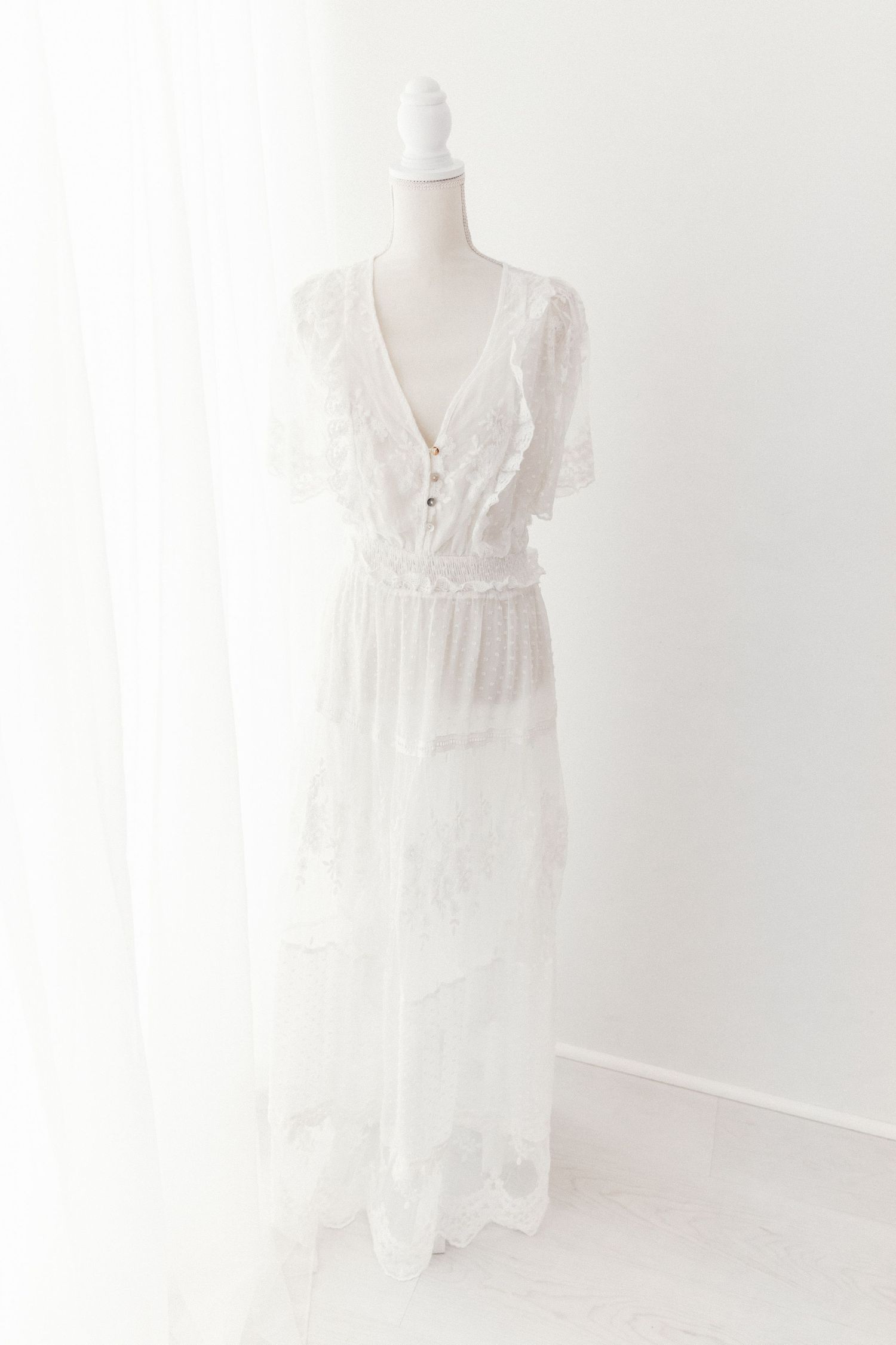 White lace maxi dress with deep V-neck, flutter sleeves, and tiered skirt on a mannequin.