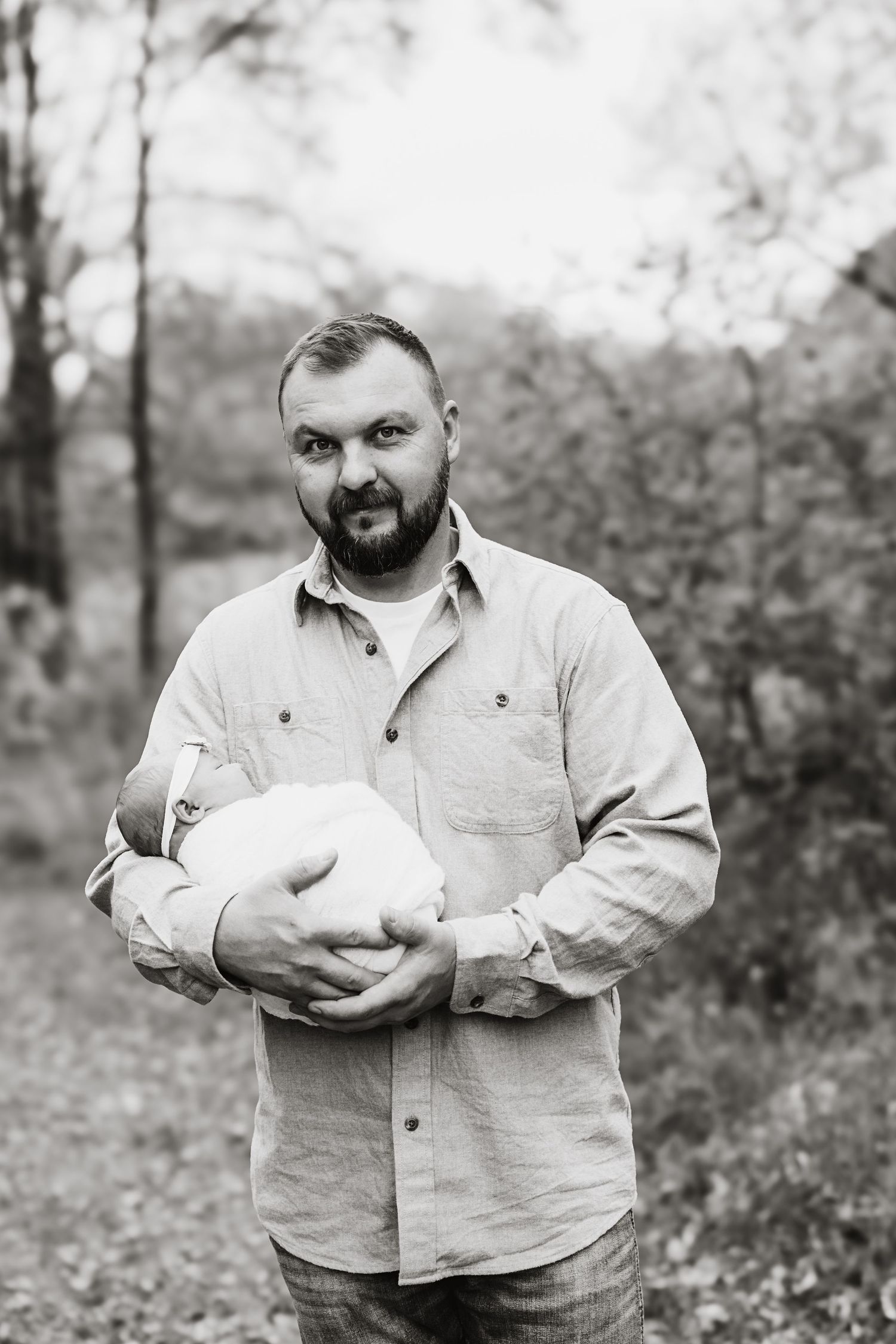 Black and white portrait of bearded father holding swaddled newborn baby in outdoor woodland setting.