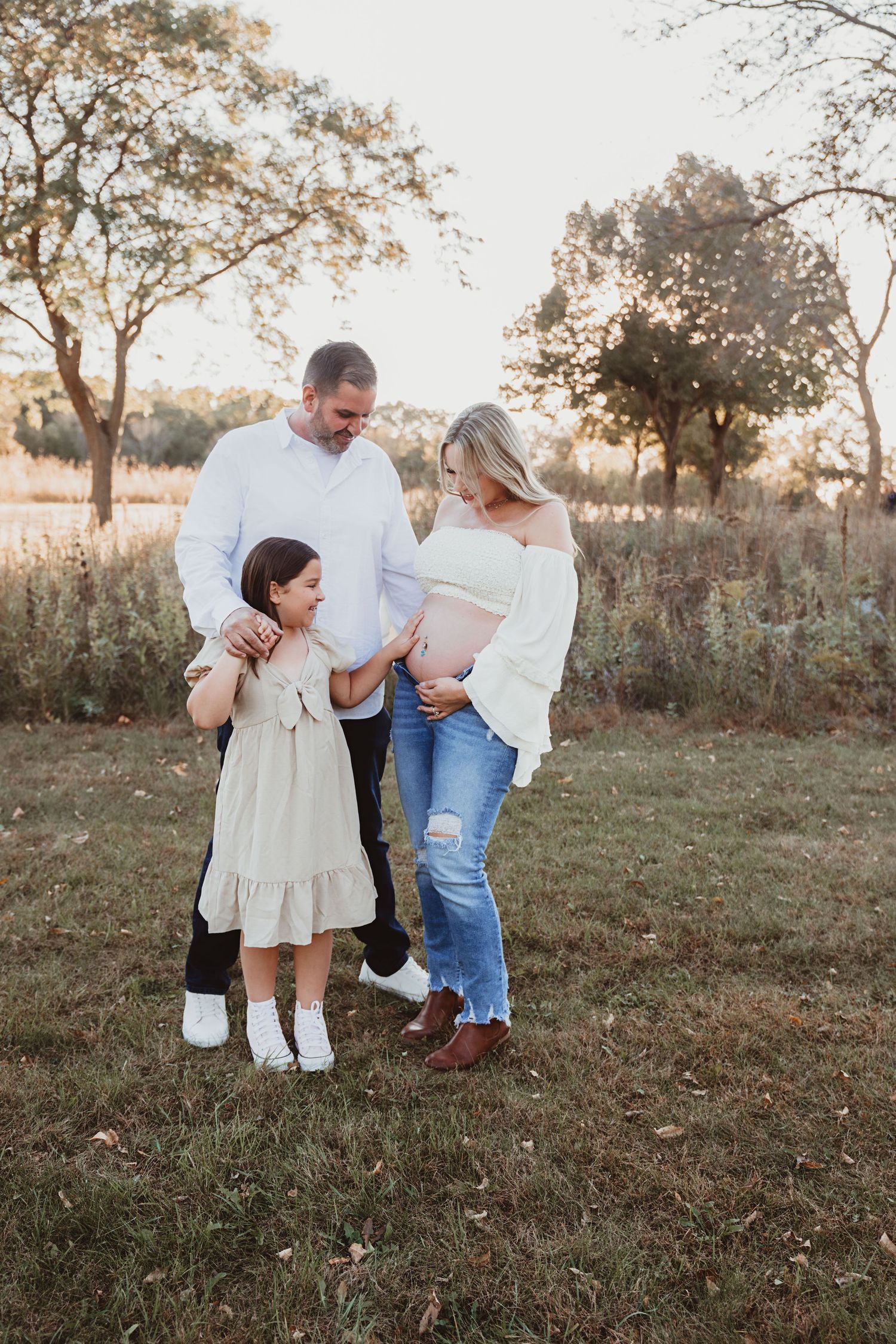 Family maternity photo outdoors at golden hour, with parents and young daughter admiring the pregnant belly.