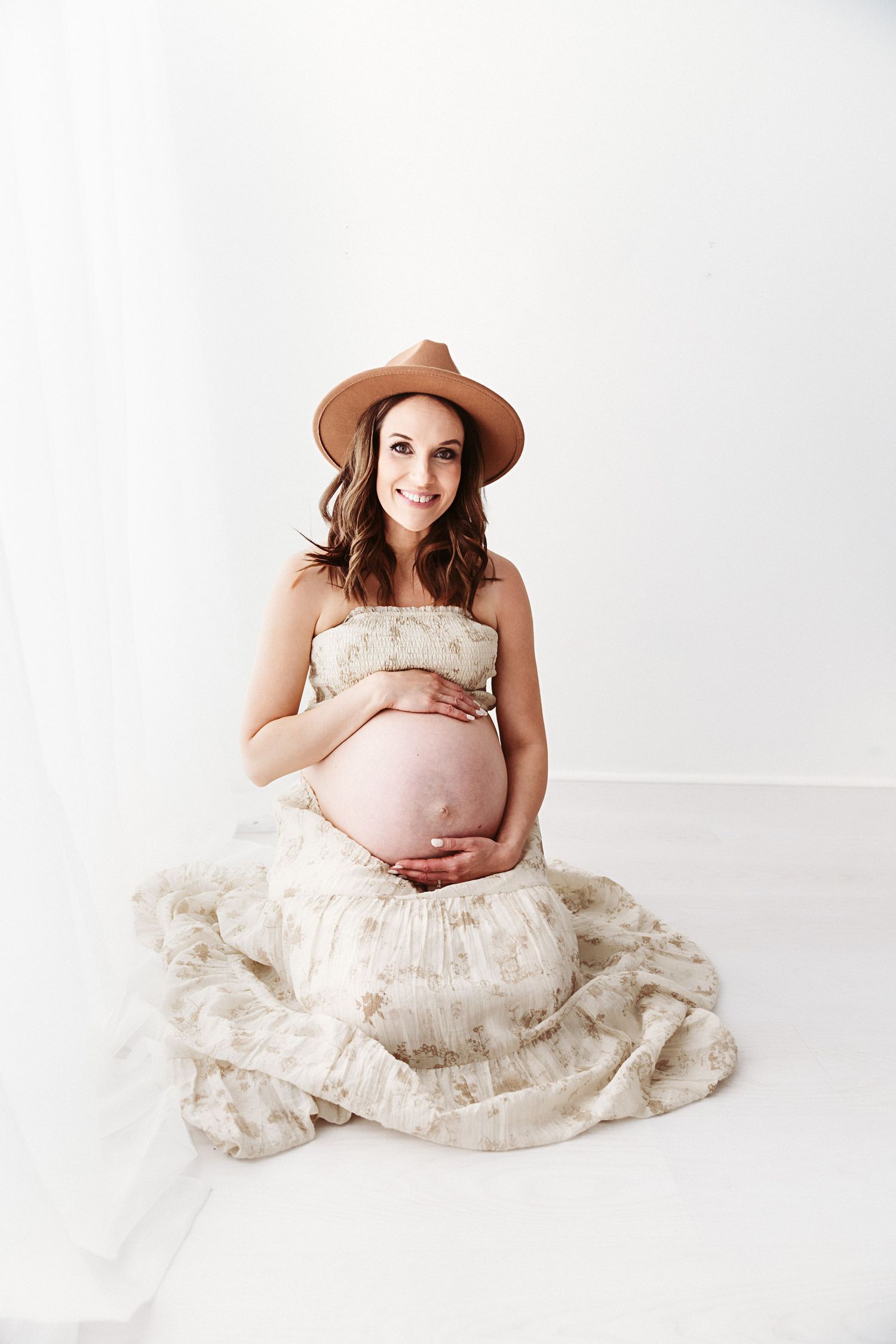 Happy pregnant woman in floral dress and brown hat sitting on floor cradling bare baby bump.