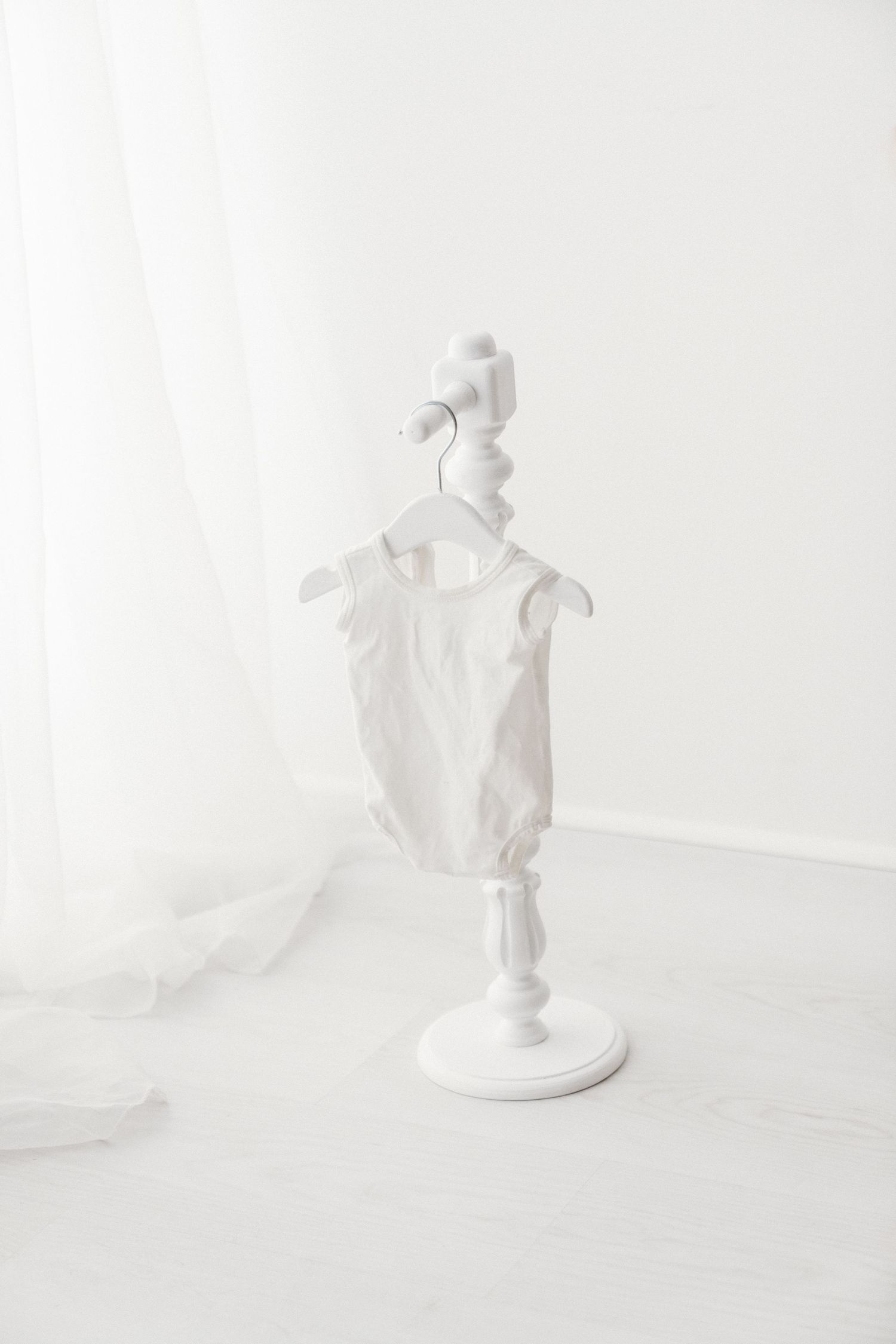 White sleeveless baby romper hanging on a white decorative hanger stand against a bright white background.