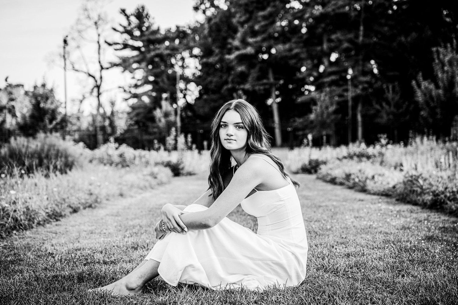 Black and white photo of a young woman in a white dress sitting gracefully on grass in a lush garden setting.
