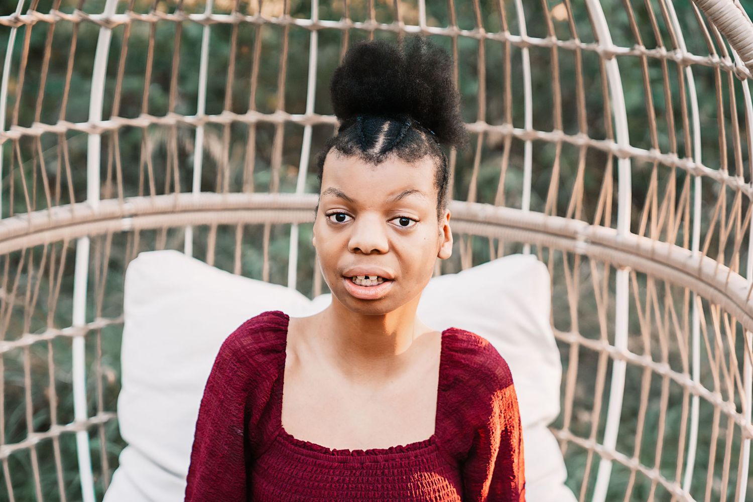 Young woman with natural updo hairstyle sitting in a wicker egg chair wearing a red smocked top.