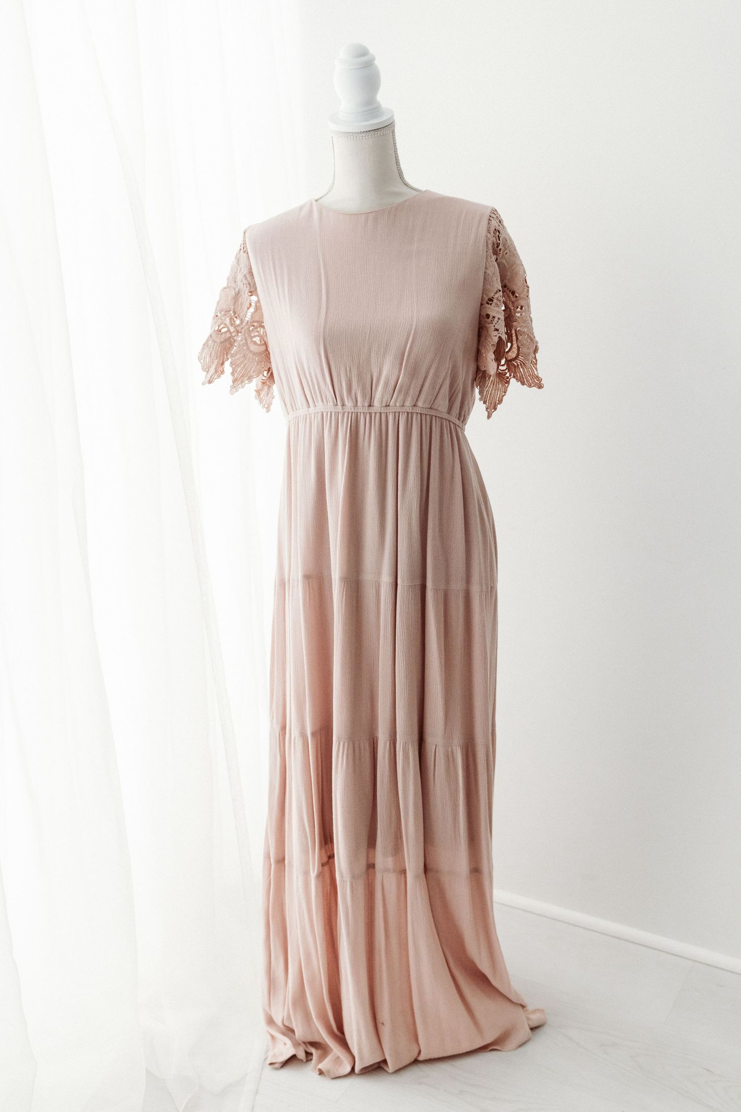 Blush pink maxi dress with lace flutter sleeves and tiered skirt on a mannequin.