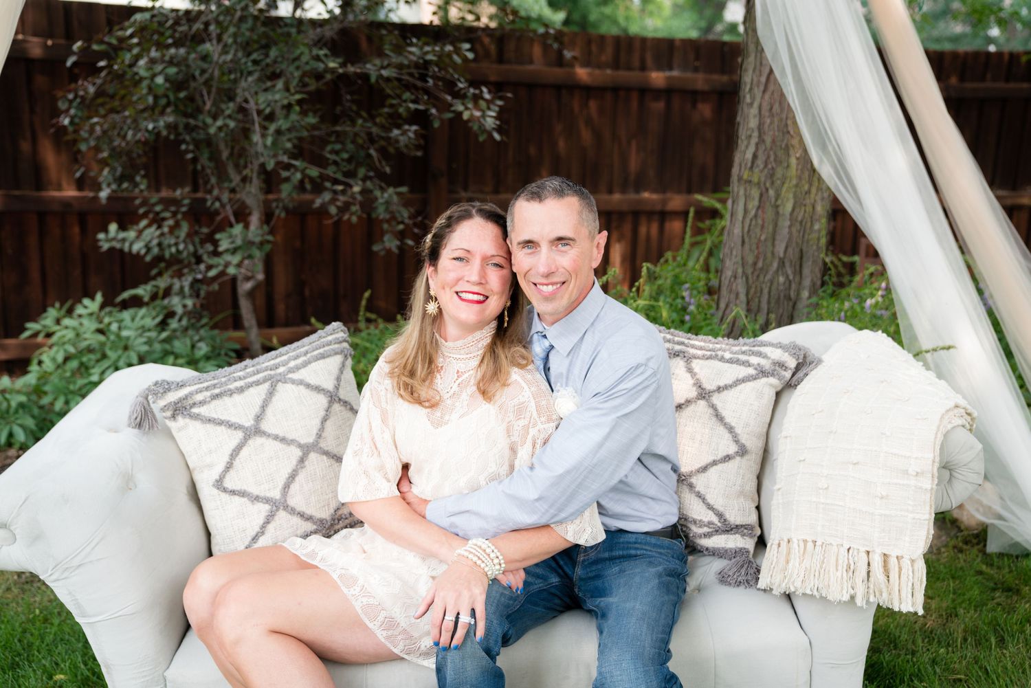 A smiling couple sits closely together on a neutral-toned outdoor sofa in a backyard setting.