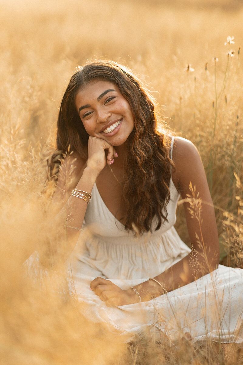 Young woman in white dress sitting in golden field, smiling warmly with hand on chin in sunlit outdoor portrait.
