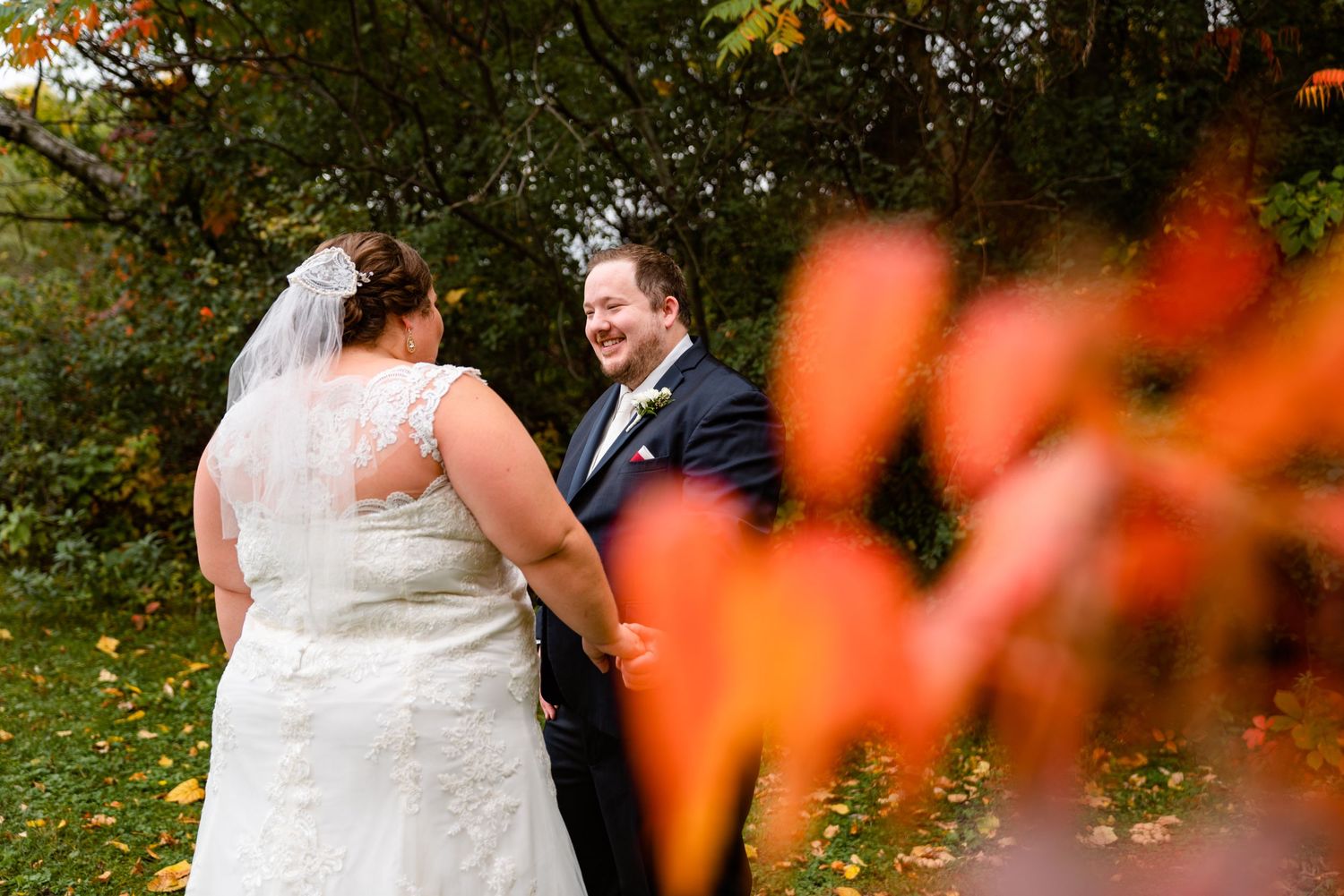 Bride and groom hold hands laughing together, framed by brilliant orange fall leaves outdoors.