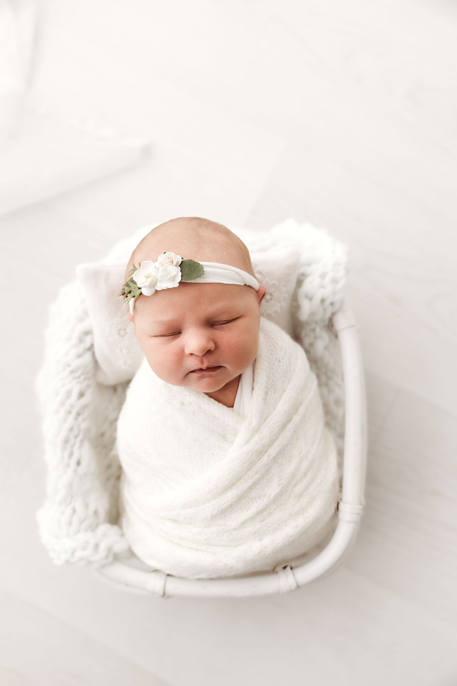 Sleeping newborn baby swaddled in white wrap with floral headband in small white bed.