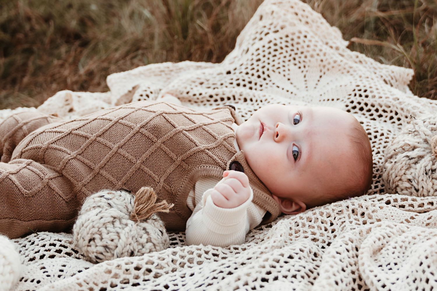 Sweet baby in diamond-knit brown romper rests on lace blanket in nature.