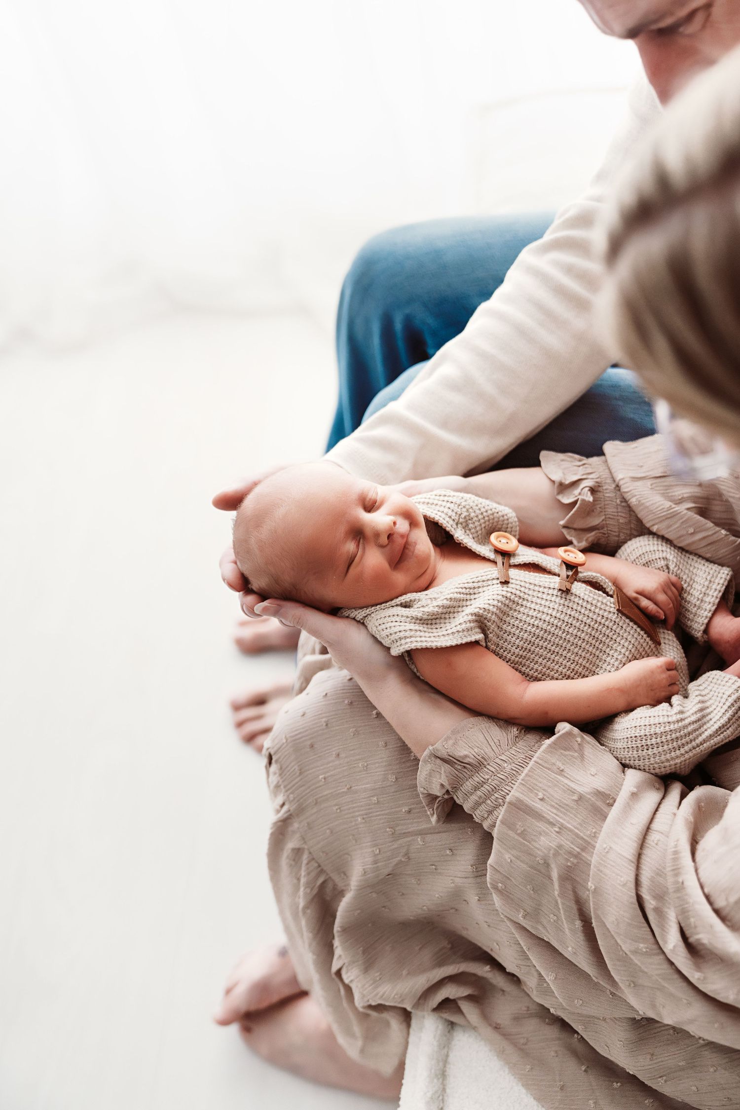 Smiling newborn baby in beige knit outfit cradled in parent's arms on soft linen blanket.