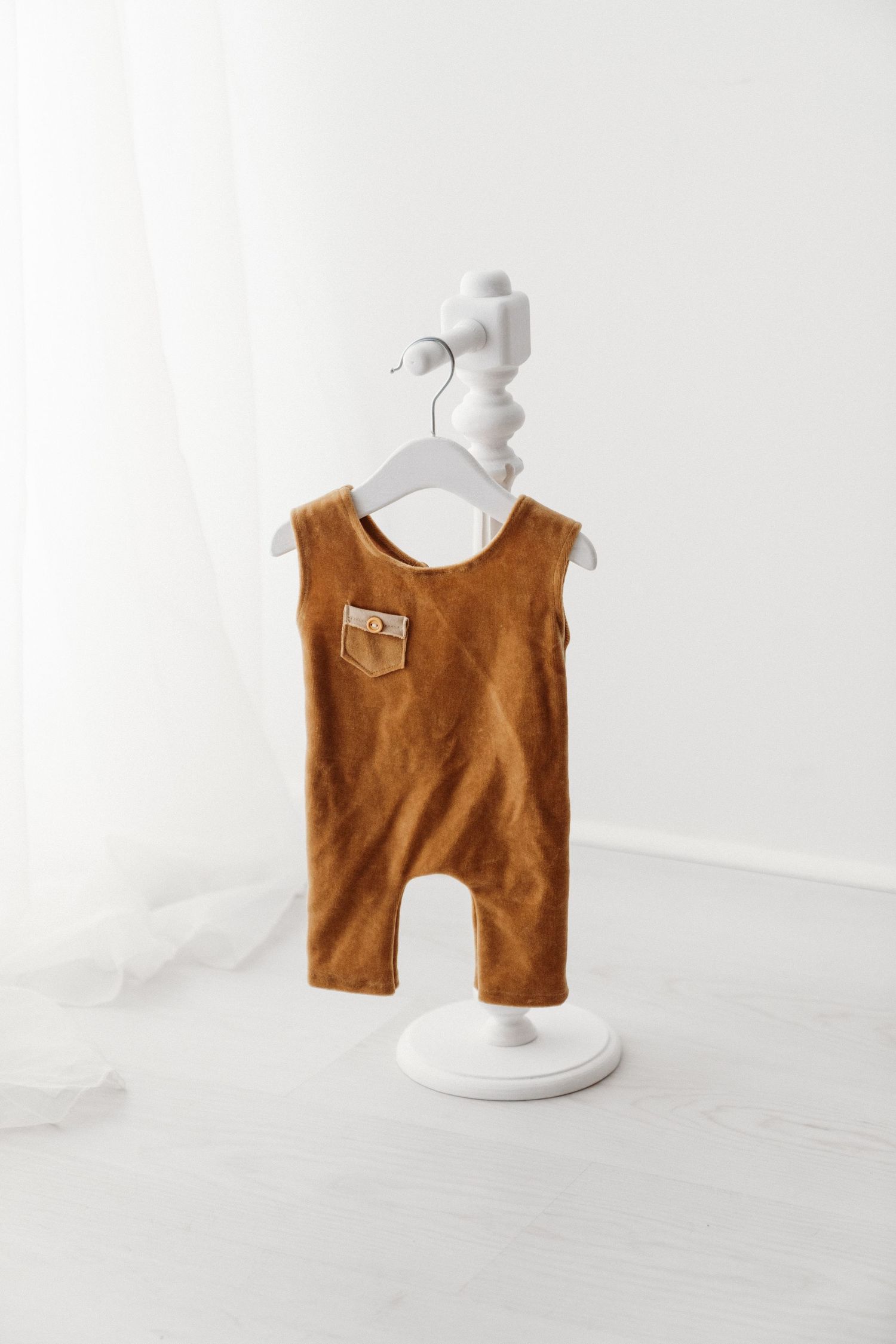 Brown suede sleeveless baby romper with chest pocket displayed on a white decorative stand.