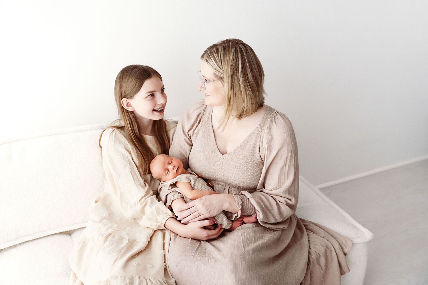 Mother and daughter smile at each other while holding newborn baby on white bed.