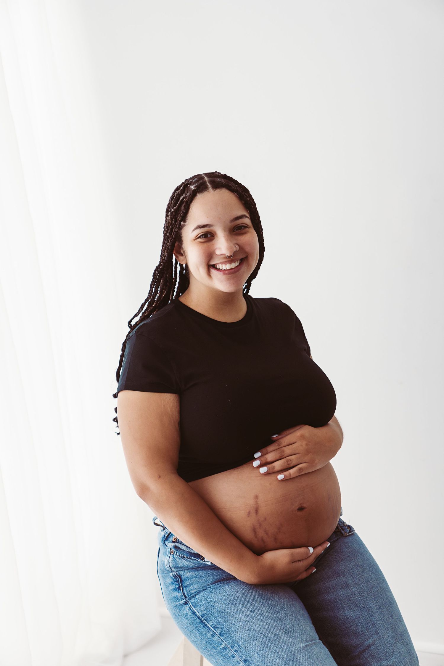 Smiling pregnant woman with braids in black crop top and jeans cradling her bare baby bump.