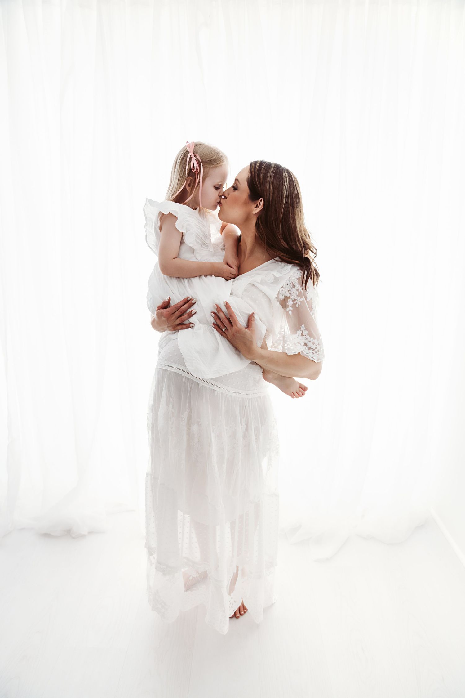 Pregnant woman in white lace gown kissing her young daughter while holding her in a studio.
