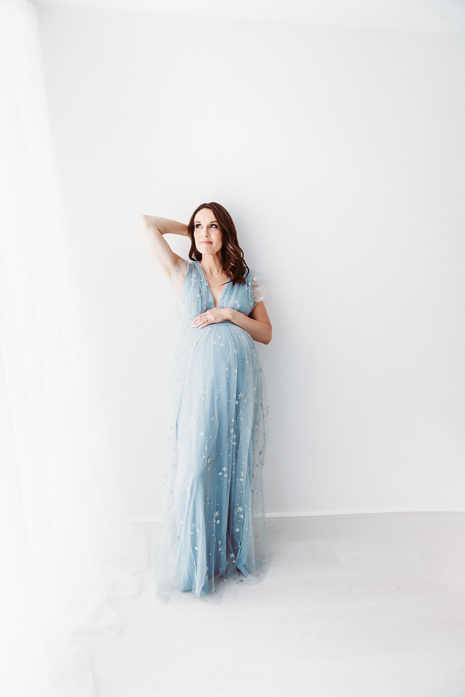 Pregnant woman in elegant blue tulle gown posing gracefully against white wall in studio maternity photo.