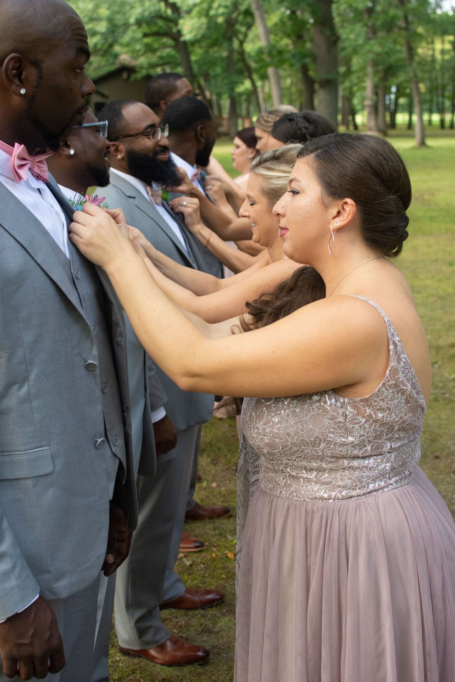 Bridesmaid in mauve dress adjusting groomsman boutonniere during outdoor wedding party photos.