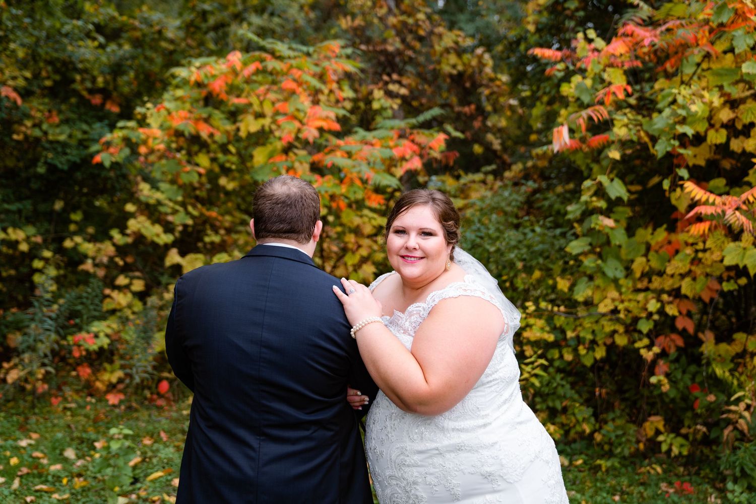 Bride smiles over shoulder at camera while embracing groom amid colorful autumn foliage backdrop.