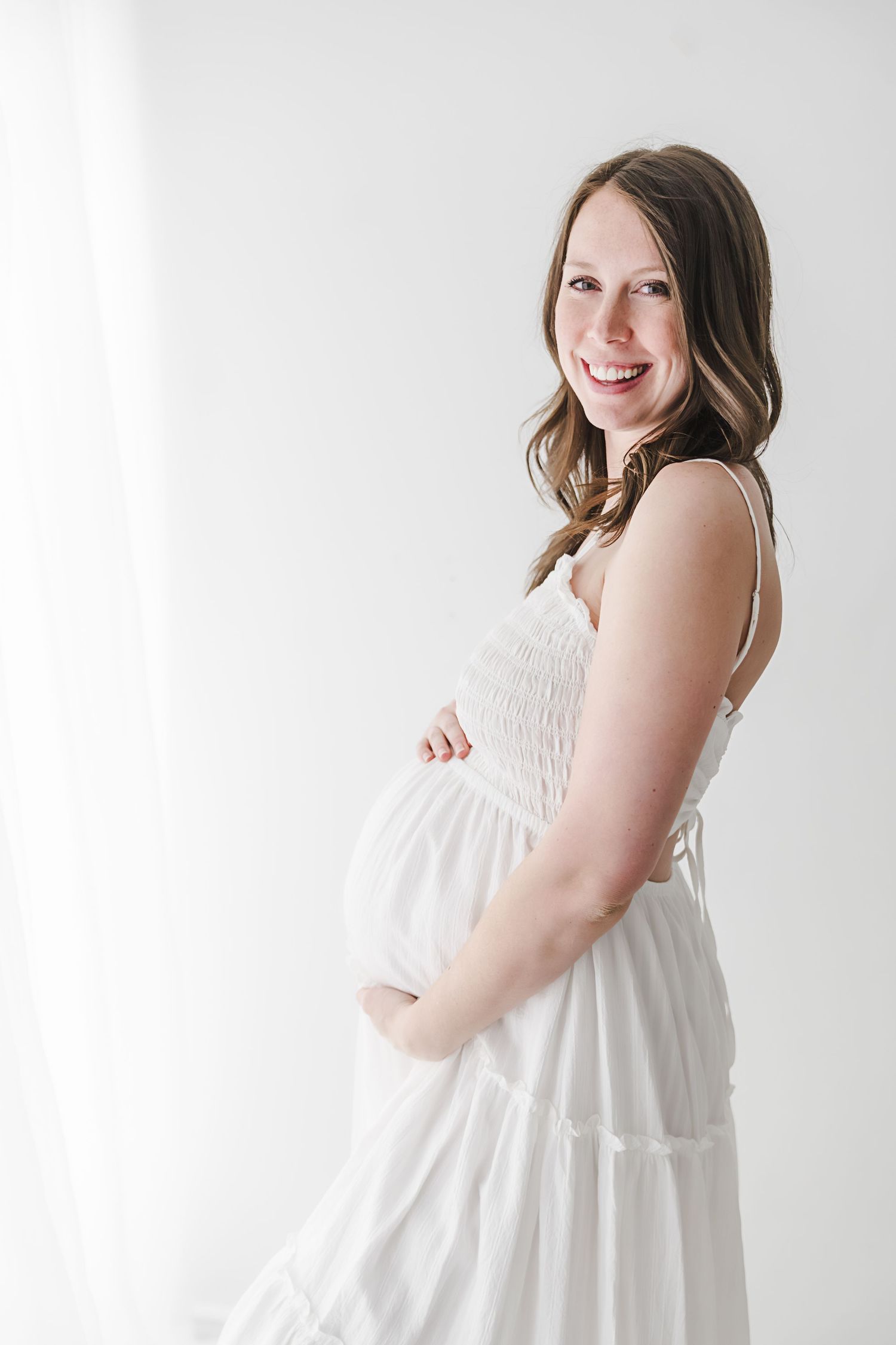 Smiling pregnant woman in white sundress cradling baby bump by sheer curtains.