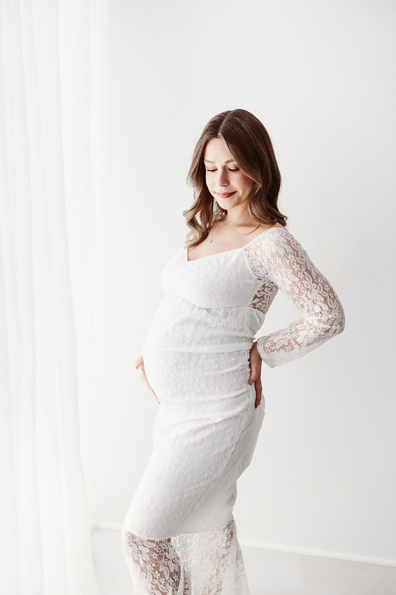 Pregnant woman in elegant white lace dress cradling her baby bump in a bright studio.